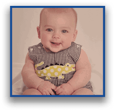 A smiling child wearing a checkered outfit with a yellow polka-dotted crocodile design is sitting on a white surface. Description generated by AI