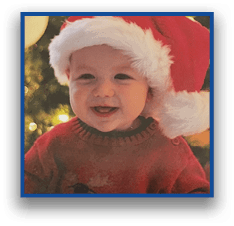 A smiling child wearing a Santa hat and a red sweater is sitting in front of a decorated Christmas tree with lights and ornaments. Description generated by AI