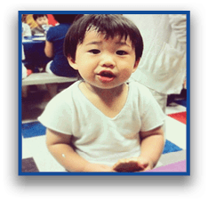A child wearing a white shirt is sitting and holding a piece of food while speaking or making a facial expression. Description generated by AI
