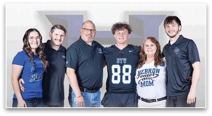 A group of six people stand closely together smiling, with one person wearing a sports jersey and another wearing a shirt that says “Hebron Senior Mom." Description generated by AI