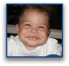 A smiling child with curly hair wearing a white shirt is looking directly at the camera. Description generated by AI