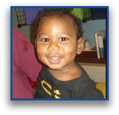 A smiling child with short curly hair is in an indoor setting with colorful storage bins and books in the background. Description generated by AI