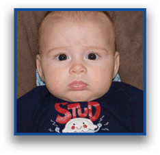 A child with short hair wearing a navy blue shirt with a cartoon character and the word “STUD" looks directly at the camera while seated on a brown and patterned cushion. Description generated by AI