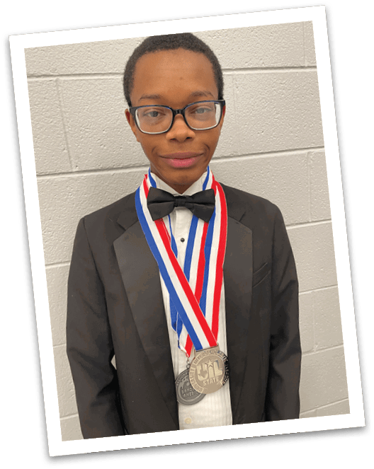 Person wearing glasses and a medal around their neck stands against a light-colored brick wall background. Description generated by AI