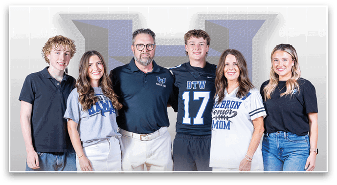 A group of six people standing closely together smiling, with some wearing casual clothing and others wearing sports-themed shirts in front of a large logo backdrop. Description generated by AI