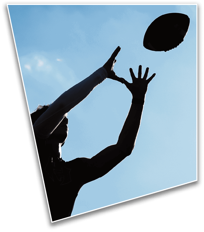 A person wearing a football helmet and gloves is holding up four fingers on one hand with a blurred football field and another player in the background. Description generated by AI