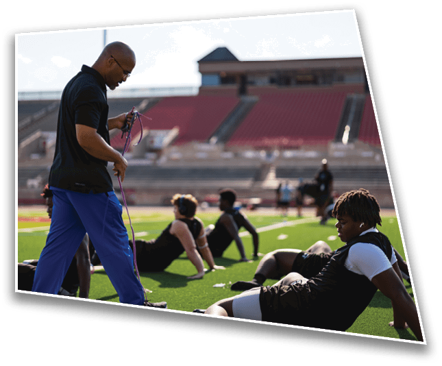 A group of people wearing football uniforms and helmets are gathered on a red field, appearing to prepare for a play or practice. Description generated by AI