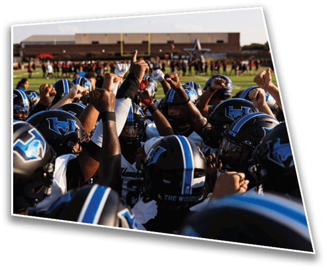 A group of people wearing football uniforms and helmets are gathered on a red field, appearing to prepare for a play or practice. Description generated by AI