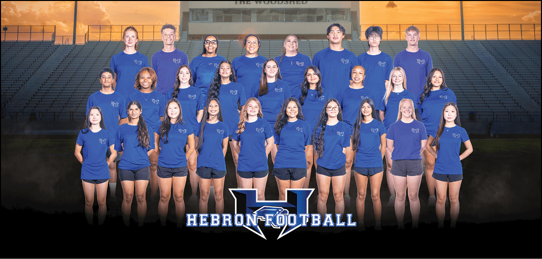 A group of people wearing matching blue shirts with a logo are standing and sitting in rows inside a large indoor space with bleachers in the background. Description generated by AI