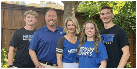 Five people wearing casual clothing with blue and black shirts featuring the word “Hebron" stand closely together outdoors in a garden setting. Description generated by AI