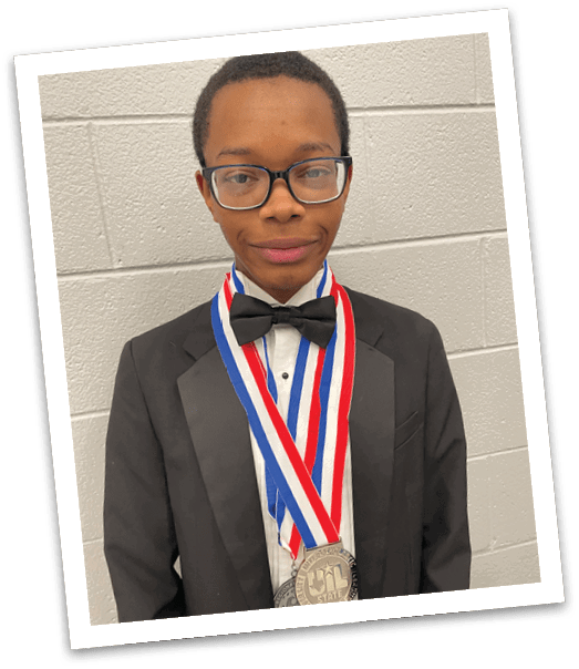 Person wearing glasses and a medal around their neck stands against a light-colored brick wall background. Description generated by AI