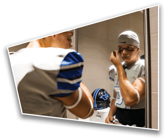 A person wearing a sports uniform and a headband is applying face paint while looking into a mirror with a football helmet visible in the background. Description generated by AI