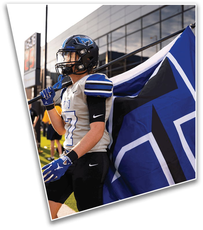 A person in a football uniform and helmet is holding a large blue and white flag while standing on a field near a building with glass windows. Description generated by AI
