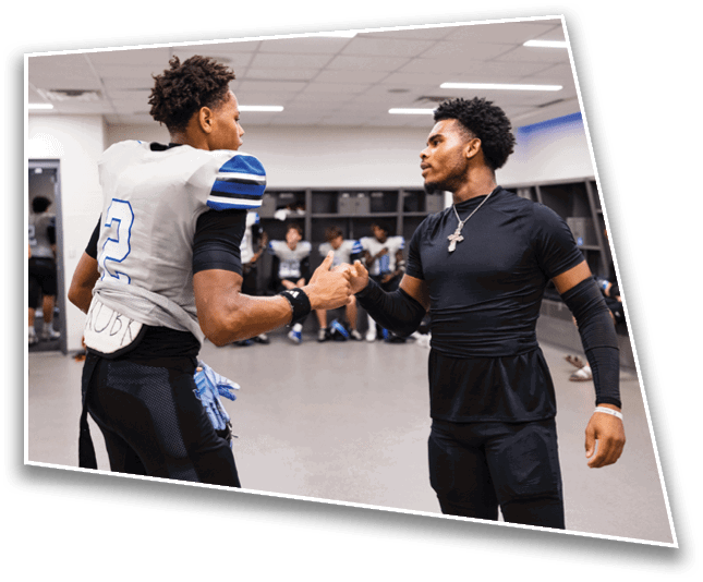 Two people in athletic clothing are fist bumping in a locker room with others sitting in the background. Description generated by AI