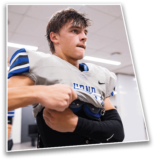 A person wearing a football uniform is being helped by another person to adjust their shoulder pads in an indoor locker room. Description generated by AI