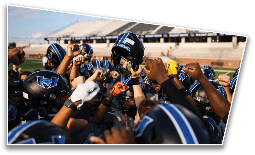 A group of football players in helmets and uniforms are gathered closely with raised fists and one player holding a helmet up in the center of a stadium field. Description generated by AI