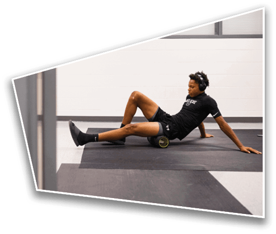 A person wearing headphones and athletic clothing is using a foam roller on their leg while sitting on a gym floor. Description generated by AI