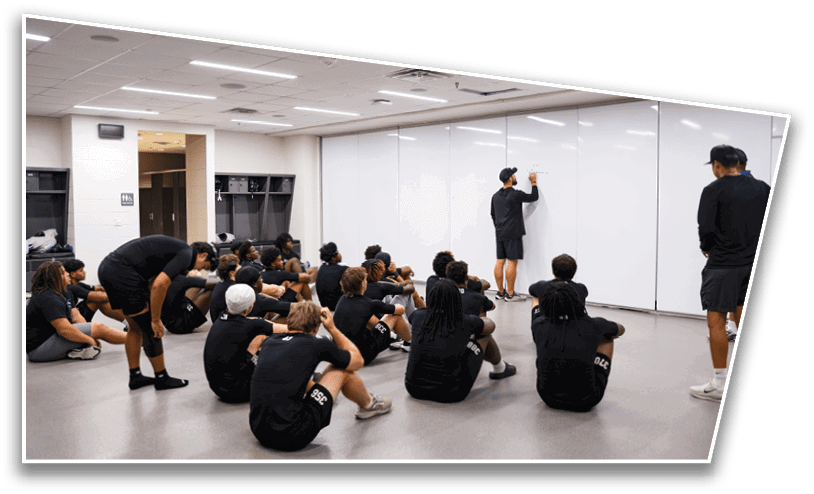 A group of people dressed in black athletic clothing are sitting on the floor attentively watching a person writing on a large whiteboard in a brightly lit room. Description generated by AI