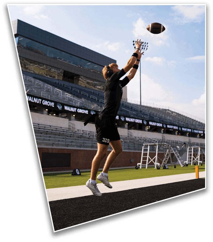 A person wearing black athletic clothing and gloves is jumping to catch a football on a sports field with empty bleachers and another person in the background. Description generated by AI