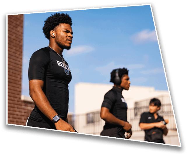Three adults wearing black athletic clothing stand outdoors with a clear blue sky in the background, one in the foreground looking focused and two others in the background, one wearing headphones. Description generated by AI