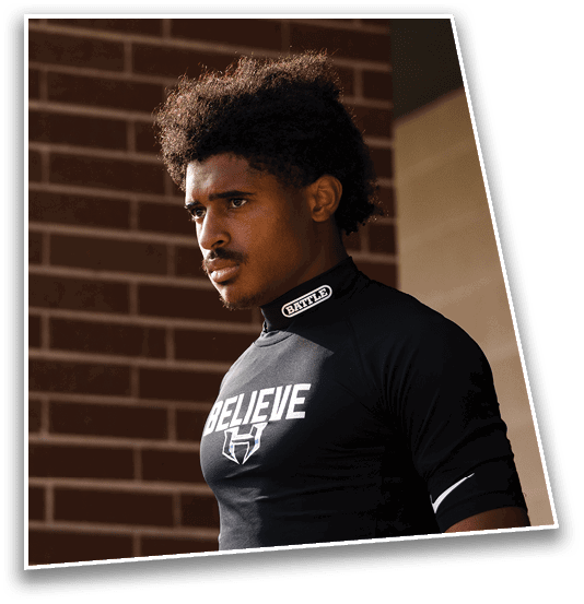 A person with curly hair wearing a black high-neck shirt with the word “BATTLE" on the collar stands in front of a brick wall looking thoughtfully to the side. Description generated by AI