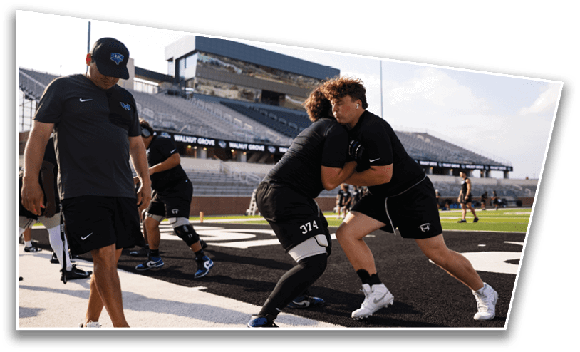 Two people in athletic clothing are engaged in a football drill on a field while a coach in a cap and sportswear observes nearby. Description generated by AI