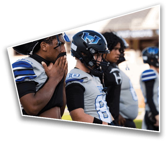 A group of people wearing football uniforms and helmets stand in a row on a field, with one person holding their face mask. Description generated by AI