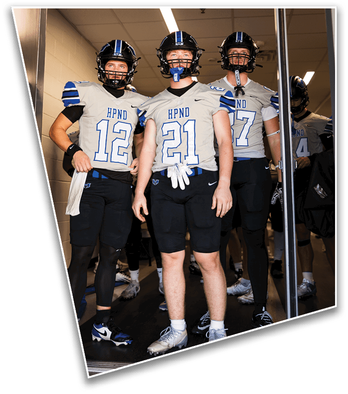 Three people wearing football helmets and gray jerseys with blue and white details stand indoors under fluorescent lighting. Description generated by AI