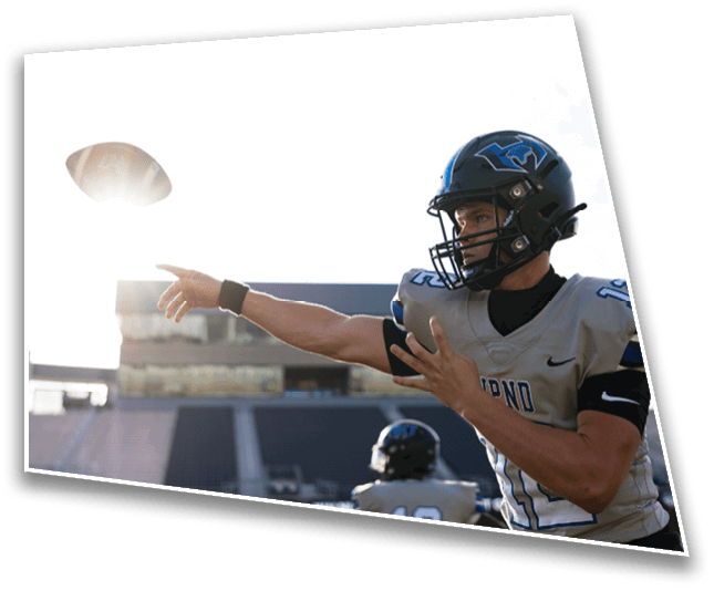 A person wearing a football helmet and uniform is throwing a football on a sunlit outdoor field with stadium seating in the background. Description generated by AI