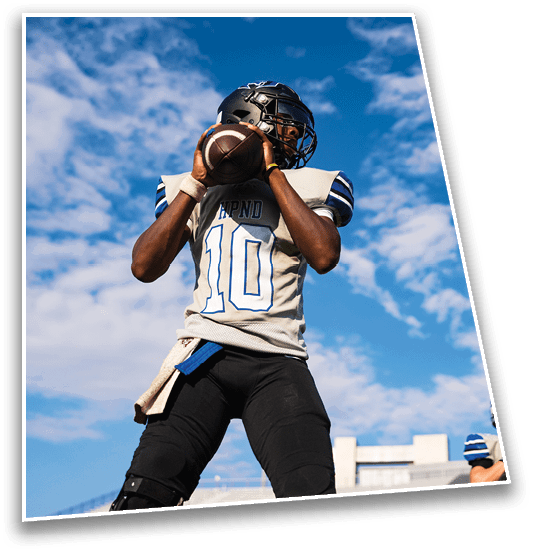 A person wearing a football uniform and helmet is holding a football in a throwing position against a blue sky with scattered clouds. Description generated by AI