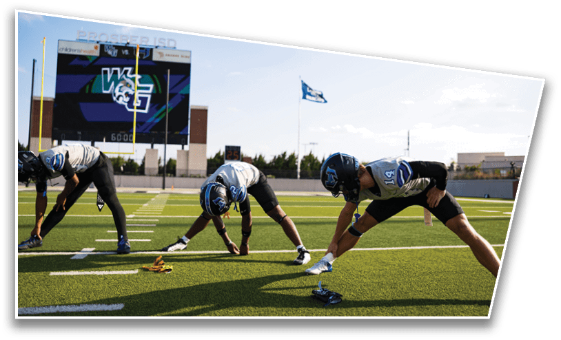 Three people wearing football helmets and uniforms are stretching on a green football field with a scoreboard and flag visible in the background. Description generated by AI