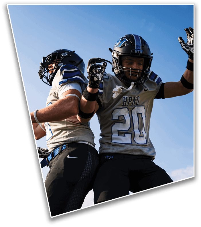 Two people wearing football helmets and uniforms are celebrating by jumping and bumping their backs against each other under a clear blue sky. Description generated by AI