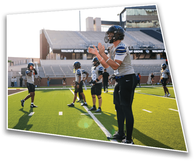 People wearing football uniforms and helmets stand on a sunlit field near the end zone with a stadium and bleachers in the background. Description generated by AI