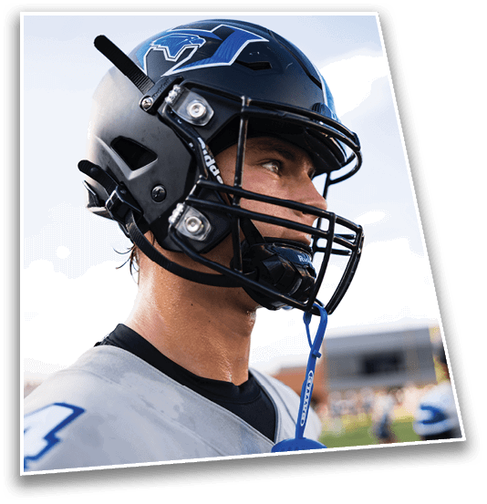 A person wearing a football helmet and jersey is looking to the side on a field with another player blurred in the background. Description generated by AI