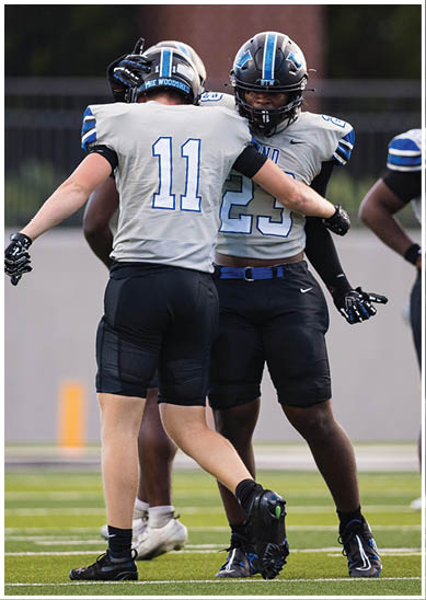 Two people wearing football uniforms and helmets are embracing on a field, with one jersey displaying the number 11 and the other 23. Description generated by AI
