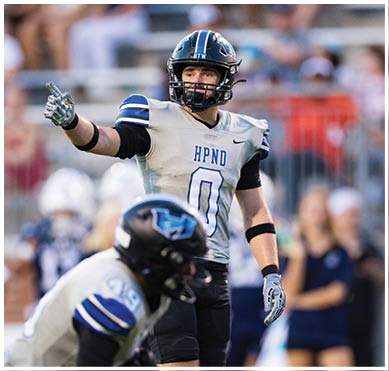 A person wearing a football uniform and helmet is pointing while standing on a field with another player in the foreground and blurred spectators in the background. Description generated by AI