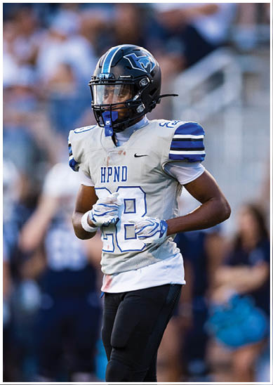 A person wearing a football uniform with a helmet and gloves is standing on a field with blurred people and bleachers in the background. Description generated by AI