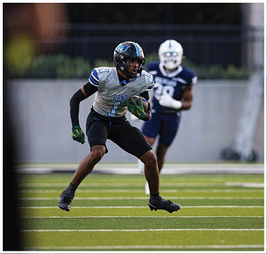 A person wearing a football uniform and helmet is running with a football on a green field while two other players in dark uniforms approach. Description generated by AI