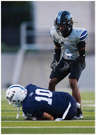 A person wearing a blue football uniform with the number 10 is lying on the ground holding a football while another person in a gray and black uniform with the number 4 stands nearby on a football field. Description generated by AI