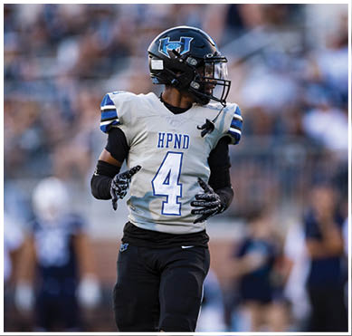 A person wearing a football uniform with the number 4 and a helmet is standing on a field with a blurred crowd in the background. Description generated by AI
