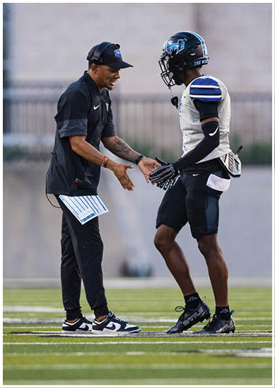 An adult wearing a black sports outfit and cap is interacting with a person in a football uniform and helmet on a field. Description generated by AI