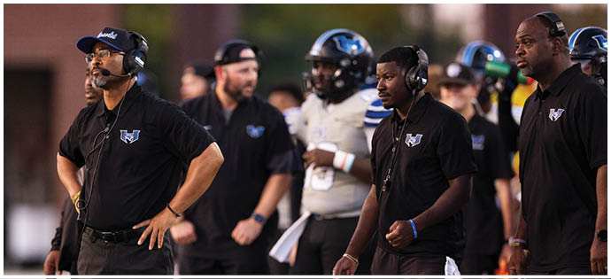 Several adults wearing black sportswear stand on the sidelines of a football field, with one person in a white uniform visible in the background. Description generated by AI