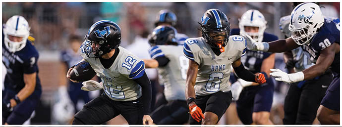 People wearing football uniforms are actively playing on a field with one person running while holding the ball and others preparing to block or tackle. Description generated by AI