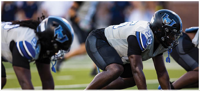 Two people in football uniforms and helmets are crouched in a ready position on a football field. Description generated by AI