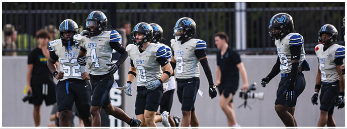 A group of people wearing football uniforms and helmets are walking on a field with two people in casual clothing in the background. Description generated by AI