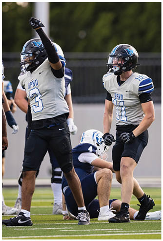 Two football players in white and black uniforms celebrate on the field while a player in a blue and white uniform sits on the ground behind them. Description generated by AI