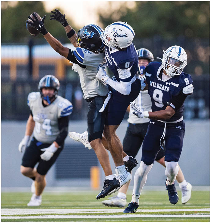 Two people in football uniforms are jumping and reaching to catch a football while two others watch nearby on a field. Description generated by AI