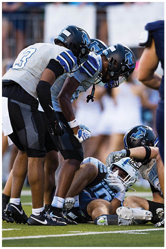 Several people wearing football helmets and uniforms are lined up in a crouched position, preparing for a play on a sports field. Description generated by AI