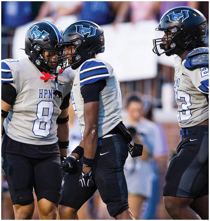 Three people wearing football uniforms and helmets are engaged in an intense moment on the field with one person clenching their fists. Description generated by AI