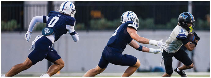 Three people in football uniforms are actively running on a field with one person holding a football while the other two appear to be attempting a tackle. Description generated by AI